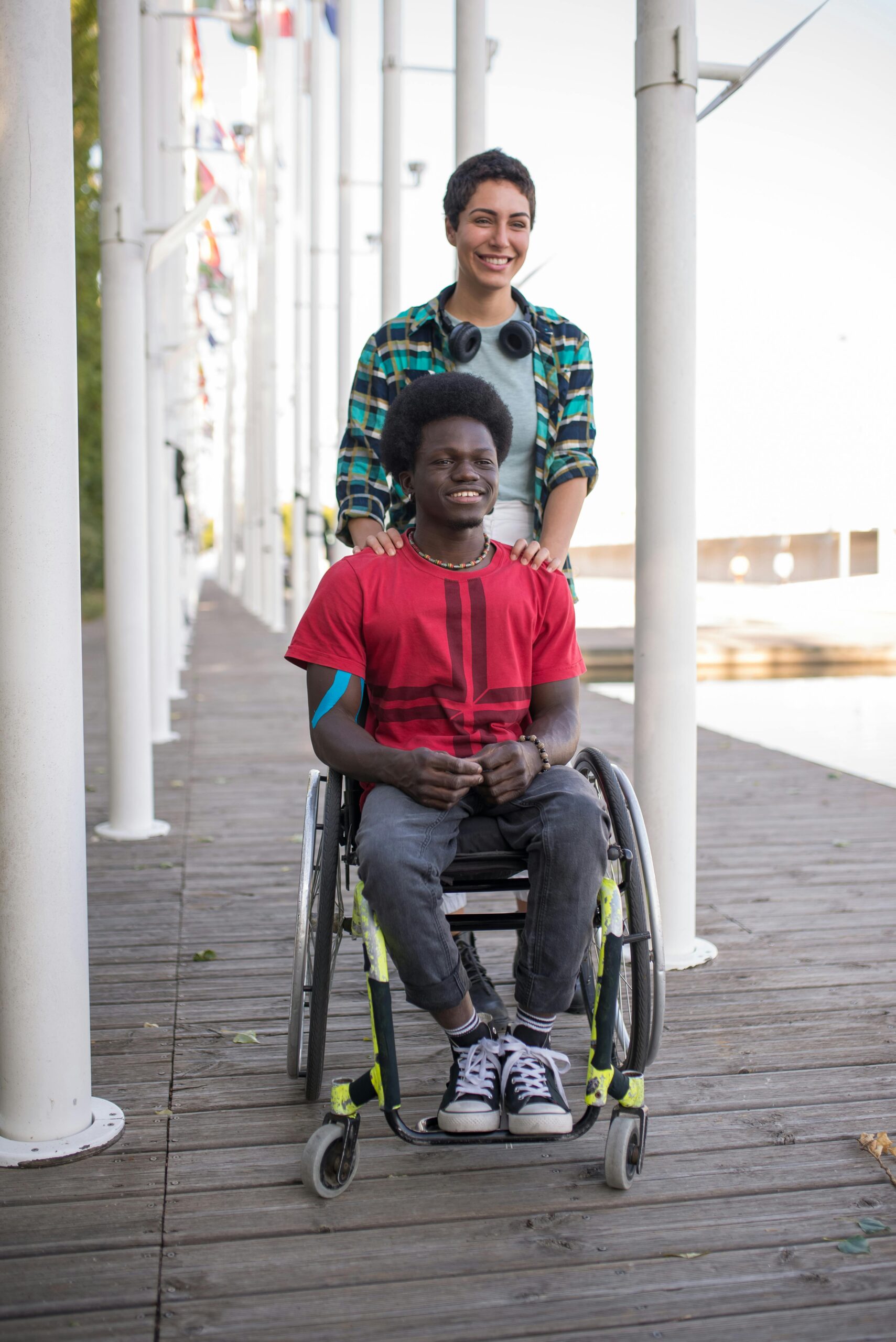 Two friends, one in a wheelchair, enjoy a laughter-filled day outdoors on a sunny boardwalk.